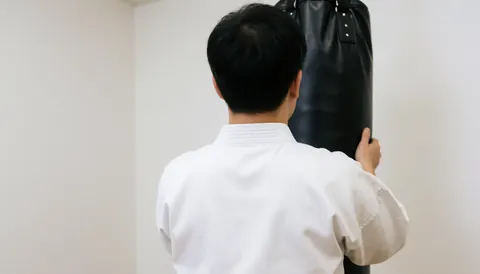 Karate student in white dobok practicing with black punching bag in dojo with white walls