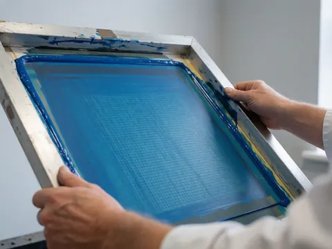Close-up of hands adjusting a mesh printing screen with blue ink for textile fabric production
