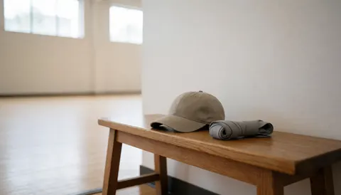 Gray baseball cap and rolled hand wraps resting on a wooden bench inside a dojo with white walls and tall windows
