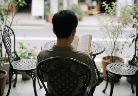 A man reads a book while seated in a wrought iron chair on a plant-filled terrace, suitable for a bed and breakfast setting.
