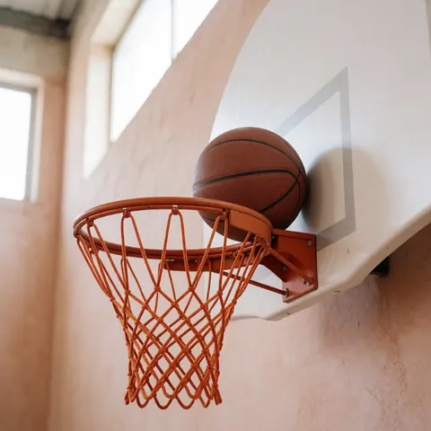 A basketball resting on the rim of a training hoop inside a gym with terracotta walls and natural light.