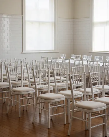 Rows of white Chiavari chairs arranged in an event room with tiled walls and a wooden floor, suitable for party coordinators.