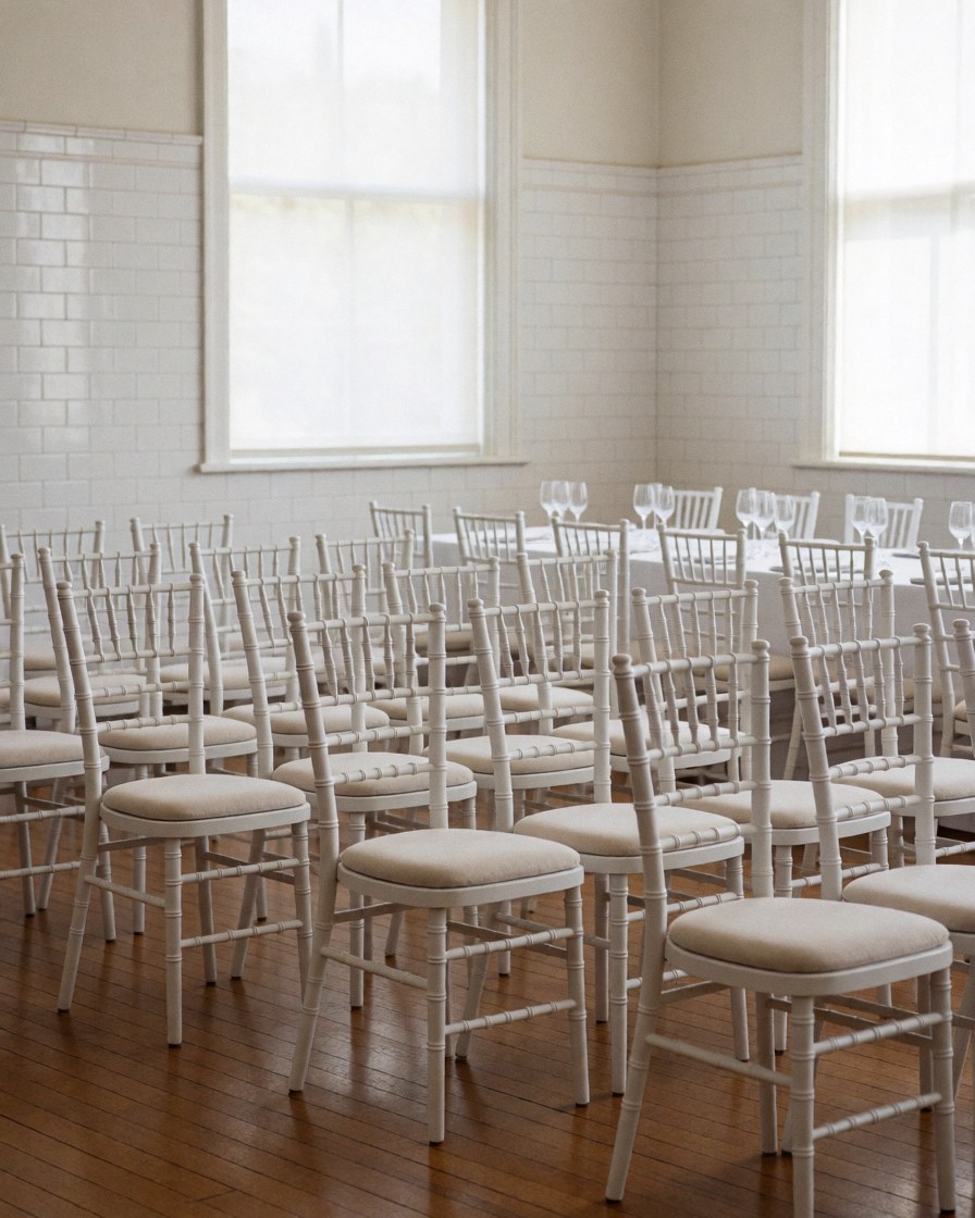 Rows of white Chiavari chairs arranged in an event room with tiled walls and a wooden floor, suitable for party coordinators.