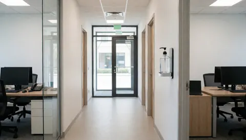Modern office corridor featuring a wall-mounted disinfectant gel dispenser, with desks, computers, and glass doors visible in the background.