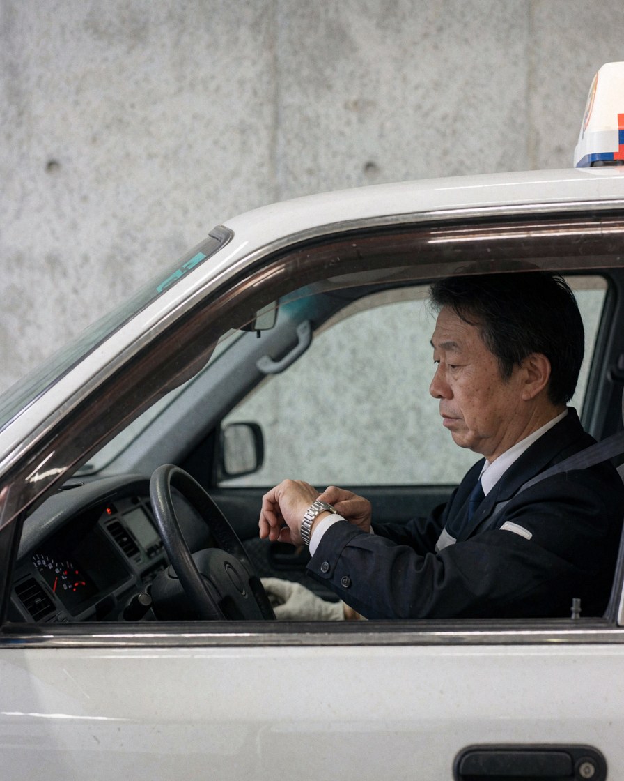 A taxi driver seated inside a parked white taxi, checking his wristwatch with concrete wall visible in the background.