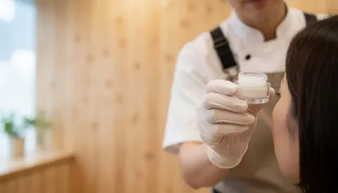 Lash technician in white glove holds gel jar near client's eye in beauty studio with light wood wall and warm lighting