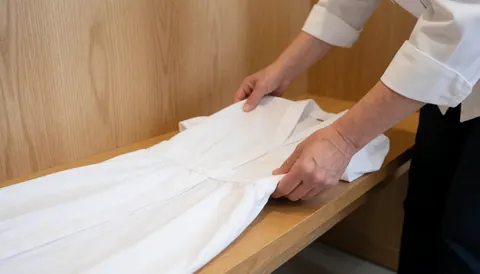 Hands folding a white garment on a light wooden surface in an ayurvedic massage setting, preparing clothing for a client.