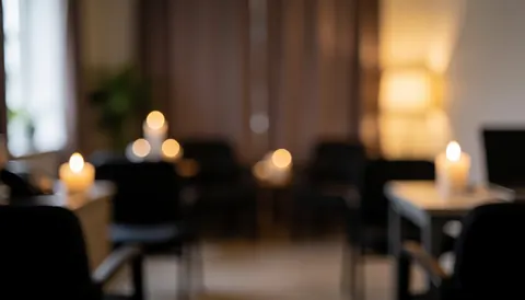 Soft focus background of a vigil room featuring lit candles, chairs, and curtains with warm, respectful lighting suitable for text overlay.