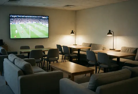 Modern interior featuring gray sofas, wooden tables, and a large wall-mounted TV displaying a soccer match, ideal for a sports club lounge.