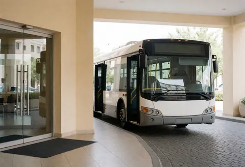 White tour bus with open doors ready to depart at the entrance of a modern hotel with beige walls and stone pavement.