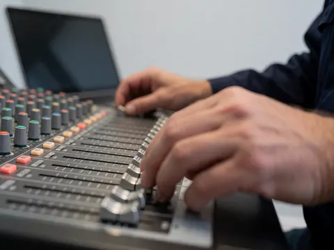 Close-up of hands adjusting faders on a professional audio mixer with a laptop and light gray wall in the background.