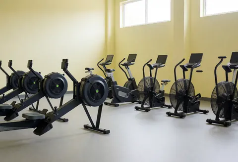 Training area featuring rowing benches and exercise bikes against light yellow walls with natural light from high windows, suitable for a CrossFit gym.