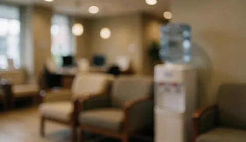 A waiting room background featuring wooden and fabric chairs, a water dispenser, and warm ambient lighting for professional care services.