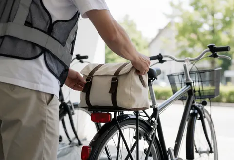 Cyclist wearing a reflective vest adjusts a fabric bag on the rear rack of a bicycle in a quiet parking area, preparing for bike taxi service.