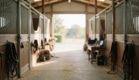A quiet stable corridor featuring hanging equestrian tack, a concrete floor, natural lighting, and a desk with a laptop for text overlay.