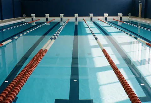 View of an empty indoor public swimming pool featuring blue walls, lane markers, and starting blocks ready for training.