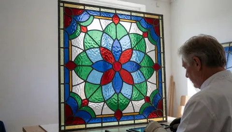 Close-up of hands assembling a floral stained glass panel with lead cames in a bright studio setting