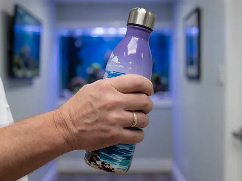 A hand holding a bottle with a marine design in an aquarium corridor with blue lighting and gray walls.