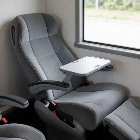 Modern public transport seat with an extended tray table, gray walls, and a window with natural light.