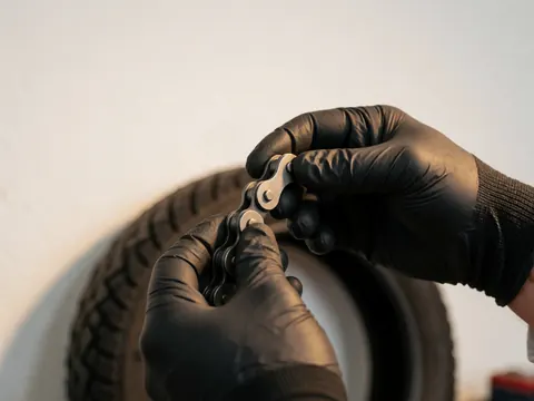 Gloved hands tightening a motorcycle chain in a workshop with a tire and white wall in the background