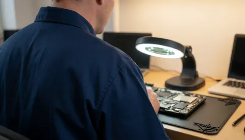 Computer repair technician working on a motherboard using a magnifying glass on an anti-static workbench.