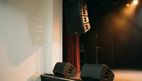 Suspended speaker array and floor monitors on a stage with warm lighting and red curtains for musical events
