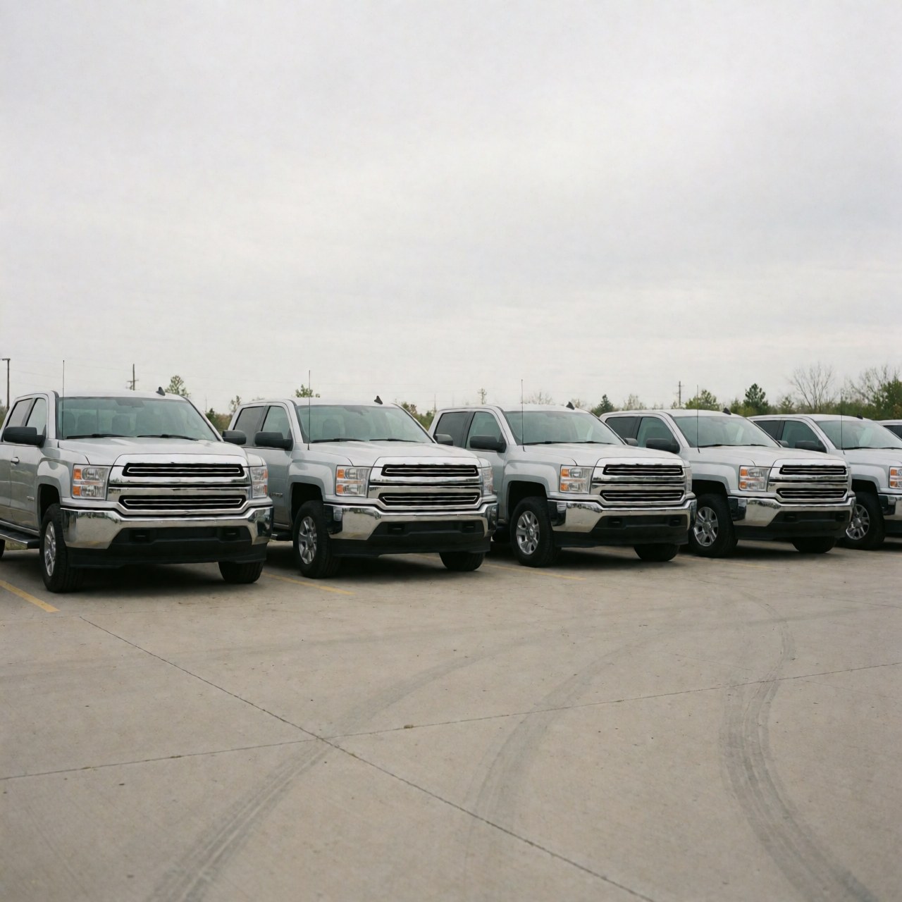 Wide view of a row of silver pickup trucks in a concrete lot under a cloudy sky, ideal for vehicle sales.