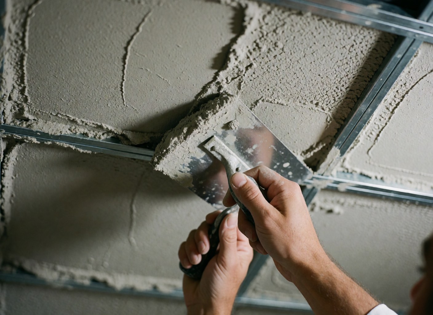 Close-up of a hand using a trowel to apply gray plaster onto a metal framework, showing rough texture and directional lighting.