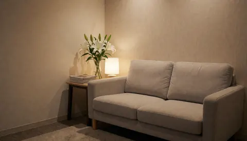 Beige sofa in a professional makeup salon waiting area with a side table holding white flowers, a lamp, textured walls, and carpeted floor