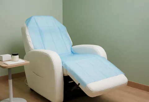 White reclining chair with sterile blue drape, side table with products, soft green walls, and light wood floor for microblading.