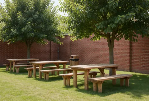Wooden picnic tables and benches under leafy trees with green grass and a red brick wall