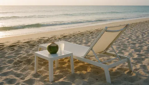 White beach chair on the sand with a coconut and a glass of coconut water, with ocean waves in the background for a beach club website hero.