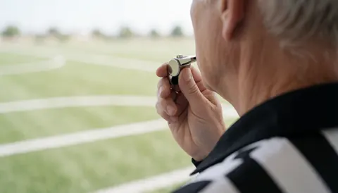 Referee blowing a whistle on a sports field during the day, viewed from behind wearing a black and white striped uniform under bright daylight.