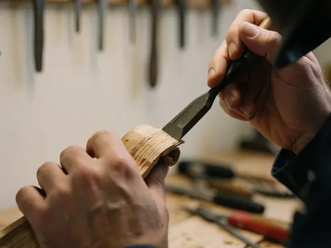 Hands using a chisel on oak wood with tools hanging in the blurred background of a carpentry workshop