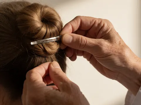 Hands adjusting a metal hair pin in a brown hair bun against a white wall, detail shot for dance instructors