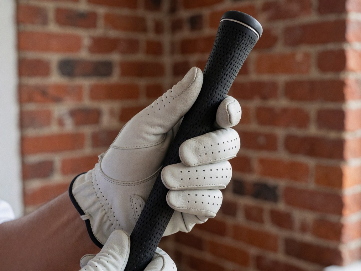 Close-up of a golfer's hand wearing a glove, holding a black perforated grip against a red brick wall, highlighting texture and tension.
