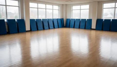Empty karate training space featuring stacked blue mats against walls and polished wooden flooring, suitable for website headers.