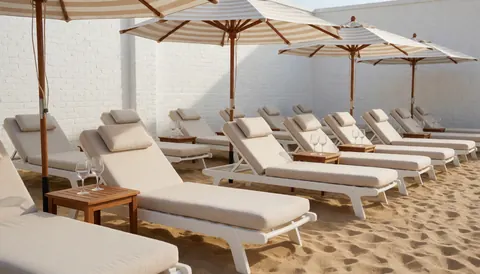 White lounge chairs with beige cushions under striped umbrellas on a beach with a white brick wall and golden sand