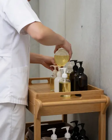 A Thai massage practitioner looks at oil dispensers on a wooden cart in a clean, professional setting.