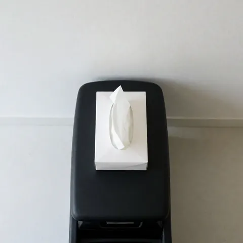 White disposable tissue box resting on a black car center console against a white wall with natural light, suitable for transportation services.
