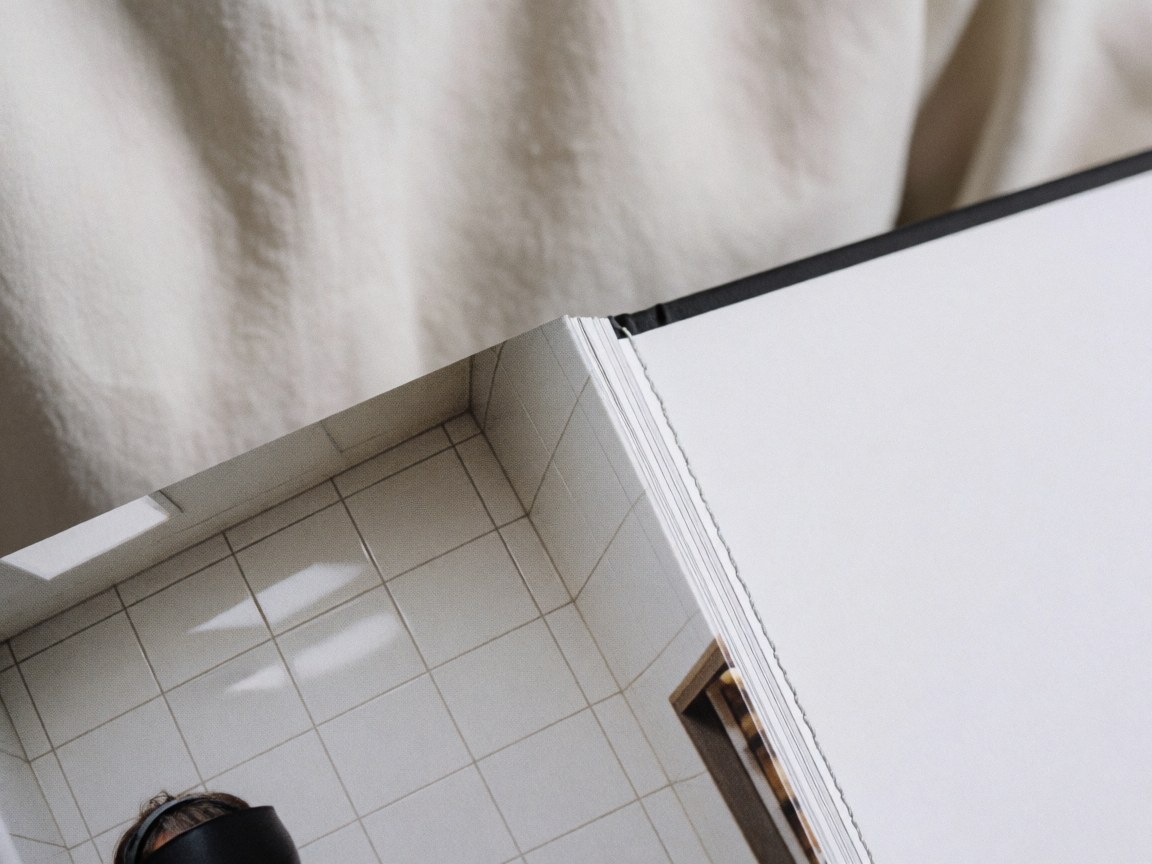 Macro detail of precise stitching on a hardcover photo album with glossy pages, set against a soft fabric background and white tiled wall.