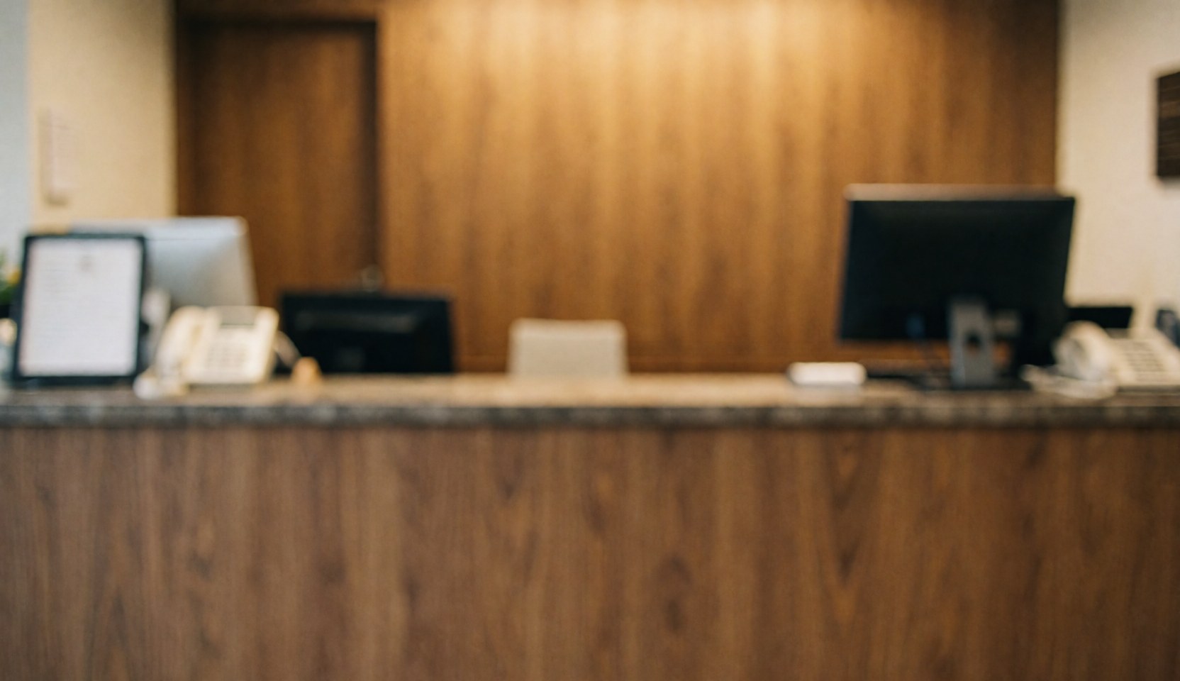 Wooden reception counter in a watch repair shop with computers and a phone, featuring warm lighting and a blurred background suitable for text overlay.
