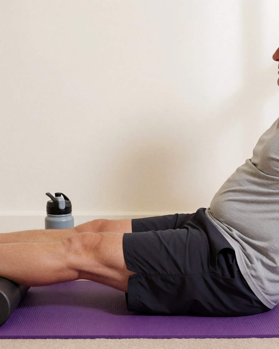 An adult performs a leg stretch using a foam roller on a purple mat near a water bottle in a rehabilitation space