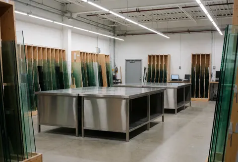 Glass workshop interior featuring stainless steel work tables, glass storage racks, and a concrete floor with LED lighting.