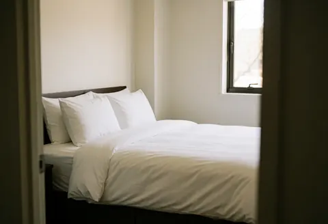 Bed with white sheets and pillows in a clean room with warm white walls and natural light from the window.