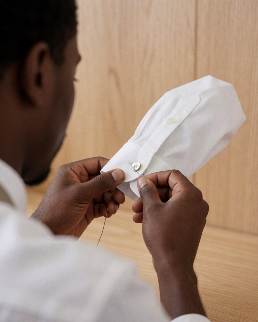 Close-up of a hand using a needle to sew a mother of pearl button onto a white shirt against a light wood background