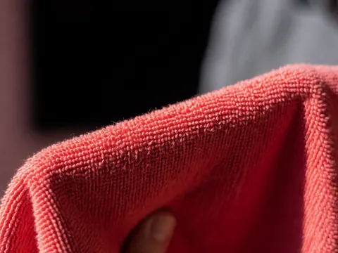 Close-up detail of a red microfiber cloth held by a hand, showing texture against a blurred black and gray background
