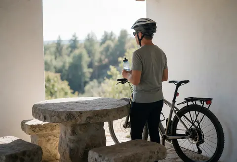 A cyclist wearing a helmet and mountain bike takes a break near a stone table at a scenic overlook with forest in the background.