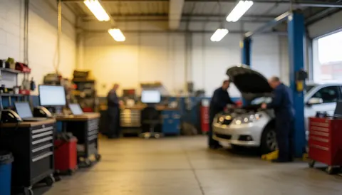 Two mechanics inspect a car with its hood open in a repair shop, surrounded by tools and industrial lights in a blurred background.