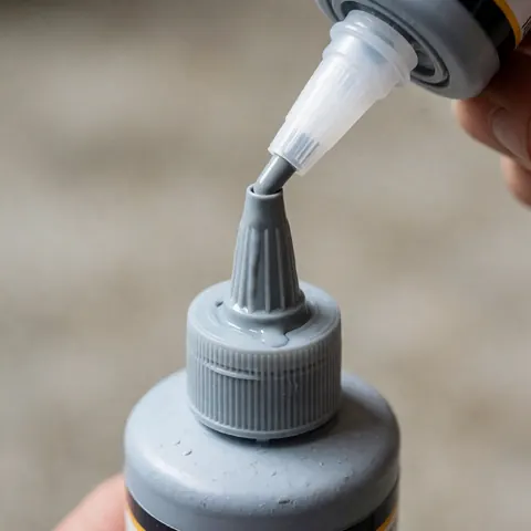 Close-up of a gray sealant dispenser with a droplet hanging from the nozzle against a concrete background, showcasing a product for bricklayers.